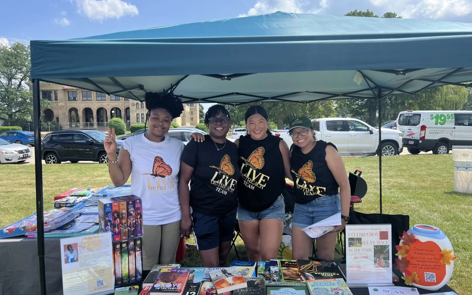 Four L.I.V.E. Outreach team members in butterfly logo shirts smiling behind a table of children's books at an outdoor community event